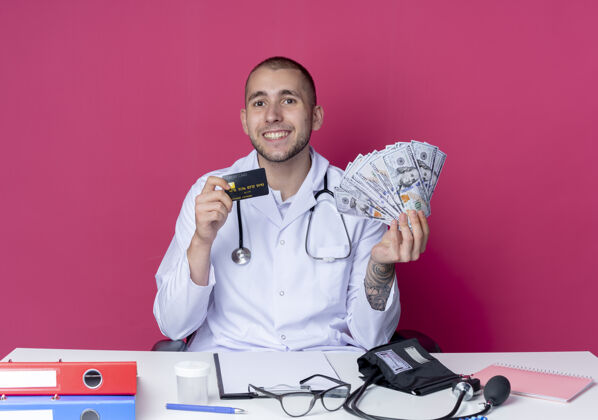 衣服面带微笑的年轻男性医生 穿着医用长袍 听诊器 坐在办公桌旁 手里拿着工作工具 手里拿着钱和信用卡 隔离在粉红色的墙上医生公民人