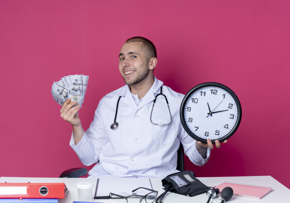 感情面带微笑的年轻男医生穿着医用长袍 听诊器坐在办公桌旁 手里拿着工作工具 把时钟和钱隔离在粉红色的墙上年轻人时钟抱着