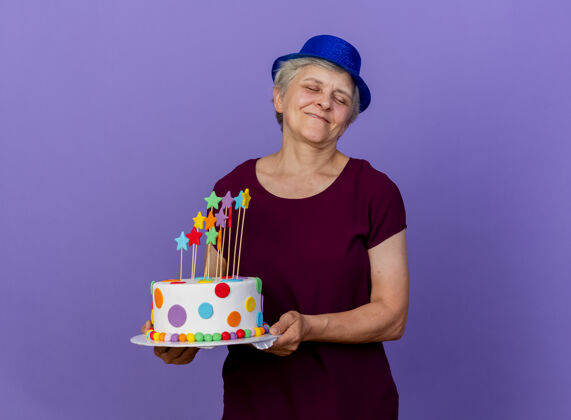 人戴着聚会帽的老太太高兴地把生日蛋糕孤立地放在紫色的墙上市民女人脸