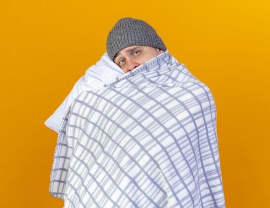金发悲伤的年轻金发病男子戴着冬天的帽子包裹在格子布举行 把头放在枕头上隔离在橙色的墙壁包裹疾病人