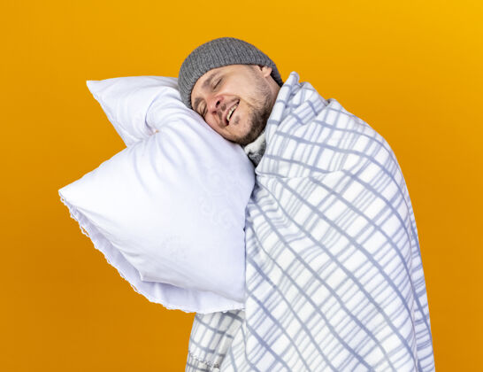 表情高兴的年轻金发病男子戴着冬天的帽子在格子布包裹举行 并把头放在枕头上隔离在橙色的墙壁人金发穿