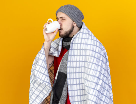 姿势一个戴着冬日帽子 围着方格围巾 喝着一杯孤立在橘色墙上的茶的年轻人包裹不愉快疾病