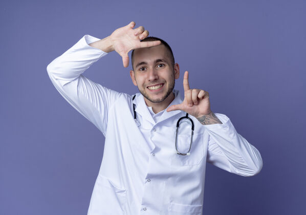 手势微笑着的年轻男医生 穿着医用长袍 脖子上戴着听诊器 在紫色的墙上做着一个孤立的姿势表情公民长袍