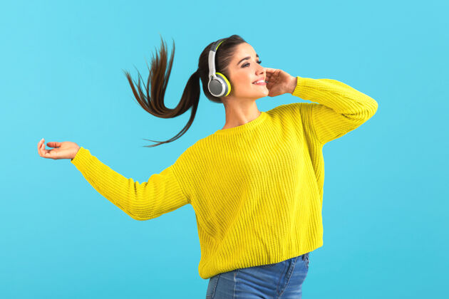 音乐戴着无线耳机听音乐的女人开心地穿着黄色针织毛衣摆着蓝色的姿势姿势欢呼无线