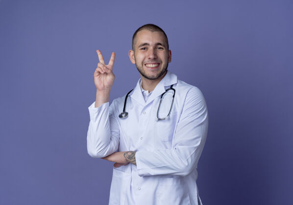 长袍面带微笑的年轻男医生穿着医用长袍 手持听诊器做着和平手势 手放在手肘下 隔离在紫色的墙上人公民男性