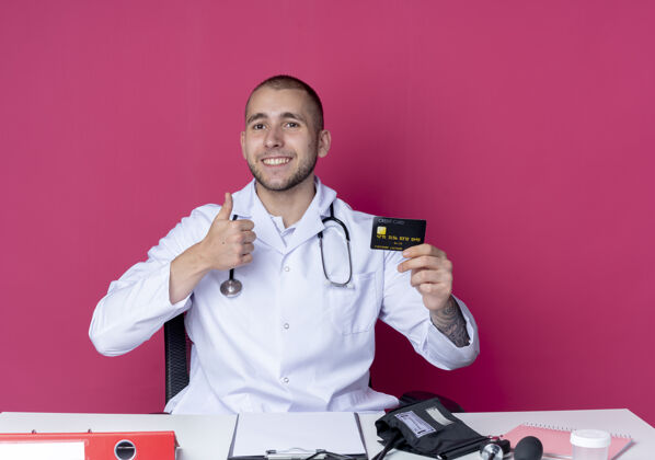 感情面带微笑的年轻男医生穿着医用长袍 听诊器坐在办公桌旁 手里拿着工作工具 手里拿着信用卡 把大拇指孤立地竖立在粉红色的墙上医生人长袍