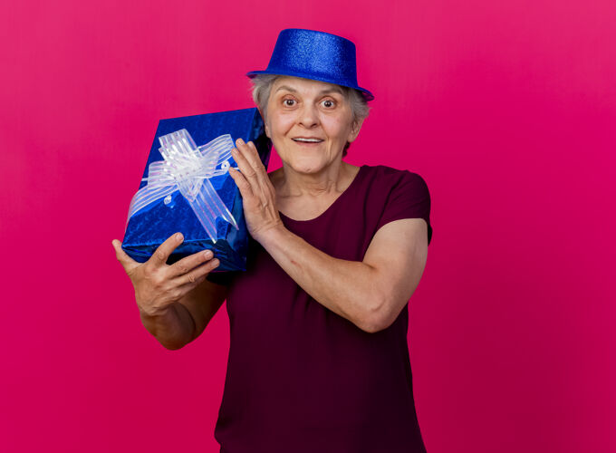 表情兴奋的老妇人戴着聚会帽 把礼品盒孤立地放在粉红色的墙上女人兴奋帽子