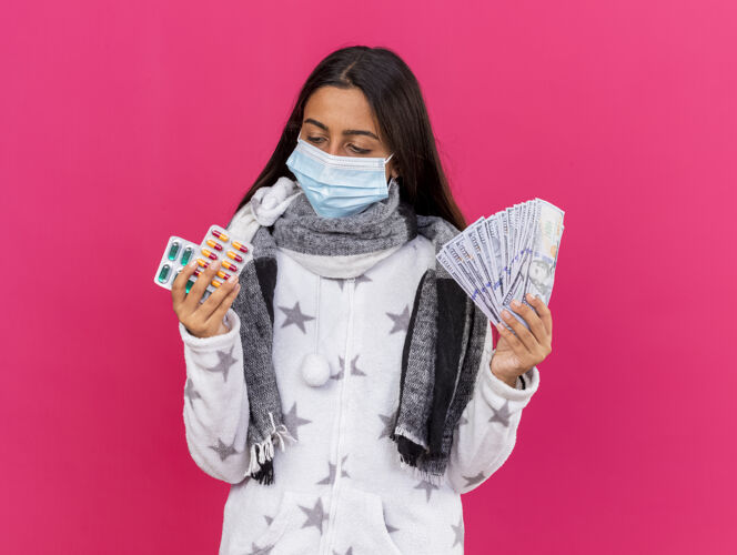 佩戴年轻的生病的女孩戴着医用口罩 戴着围巾 手里拿着钱 看着药片孤立在粉红色的手上围巾思考疾病