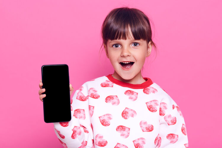 在线惊讶的小女孩拿着智能手机 一脸惊讶的表情 张开嘴 隔着粉色的墙壁摆出孤立的姿势博主联系人大笑