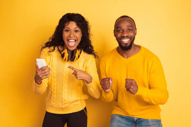 非洲年轻感性的非裔美国人 穿着黄色背景的明亮休闲服美丽的情侣人类情感的概念 面部表情 关系在智能手机上震惊地指着女孩微笑人类