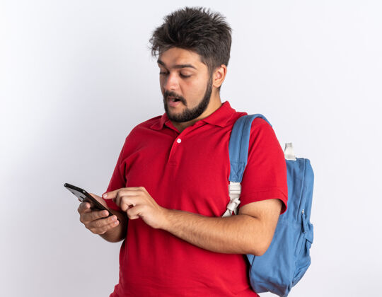 胡子快乐的年轻留着胡须的学生 穿着红色马球衫 背着背包 手持智能手机 站在白色背景上微笑着输入信息衬衫手机背包