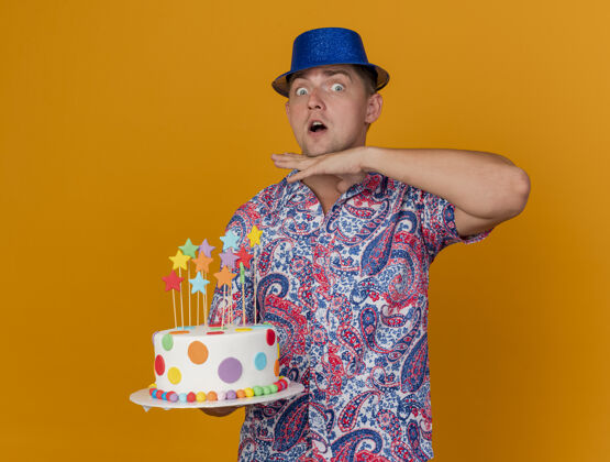 蓝色惊讶的年轻人戴着蓝色帽子拿着蛋糕 手放在喉咙上孤立的橙色蛋糕抱着小伙子