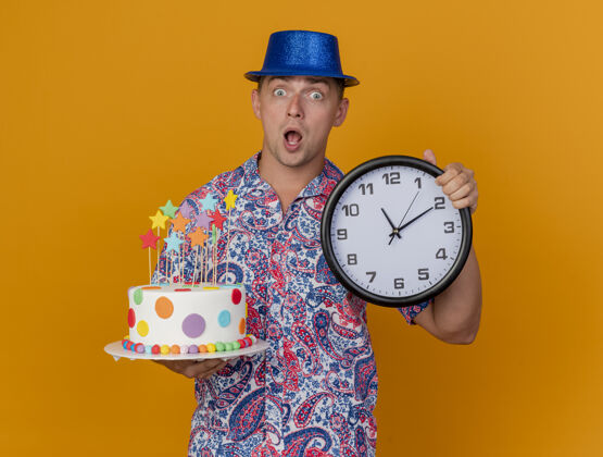 派对惊讶的年轻人戴着蓝色帽子拿着挂钟蛋糕孤立在橙色年轻人小伙子时钟