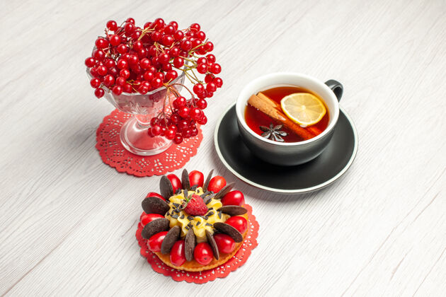花边俯视图红色椭圆形蕾丝装饰物上的水晶杯里的红醋栗 白色木桌上的柠檬肉桂茶和浆果蛋糕杯子水晶红色