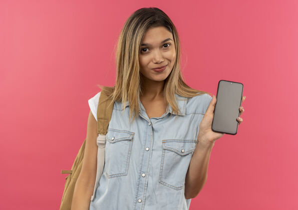电话高兴的年轻漂亮的学生女孩穿着背袋显示手机在前面隔离在粉红色的墙上请女孩人