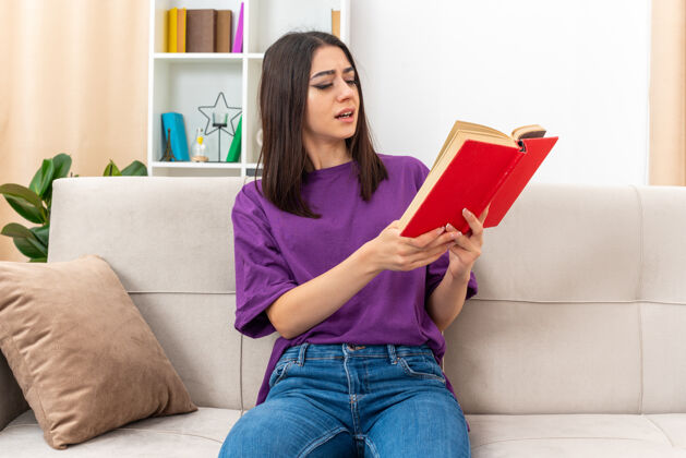 衣服一个穿着休闲服的年轻女孩拿着一本带着困惑表情的书在家里看书 坐在客厅的沙发上休闲女孩房间