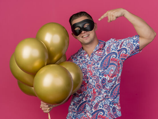 点面带微笑的年轻人戴着化装眼罩 手持并指着粉红色的气球戴着面具气球