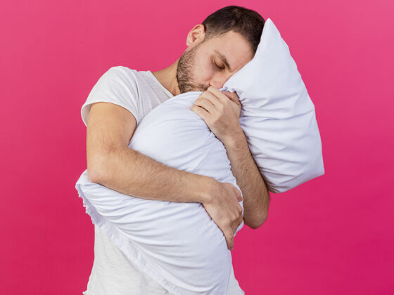 眼睛歪着头的年轻病夫抱住了孤立在粉红色枕头上的枕头男人年轻拥抱