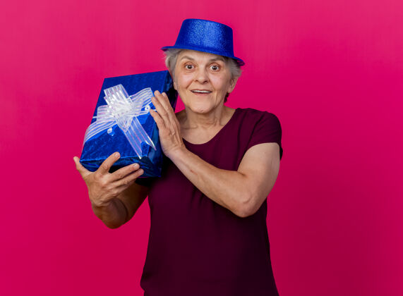 表情兴奋的老妇人戴着聚会帽 把礼品盒孤立地放在粉红色的墙上女人兴奋帽子