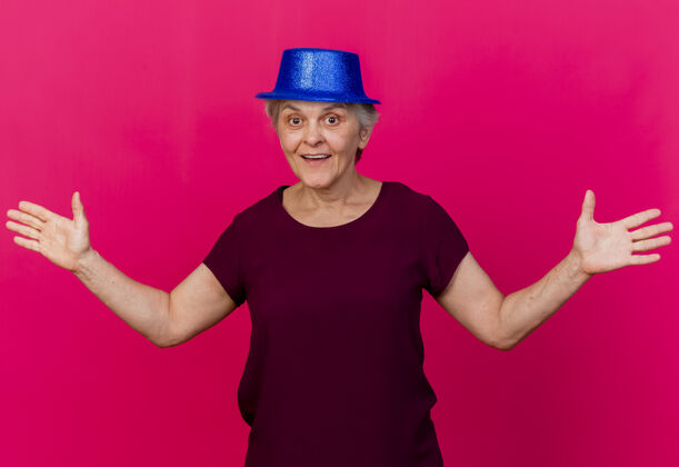 老人惊讶的老妇人戴着党的帽子站在摊开双手看着前面孤立的粉红色墙壁穿脸人