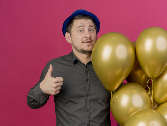 表演高兴的年轻人戴着蓝色的帽子站在气球旁边 拇指向上 孤立地站在粉红色的气球上帽子小伙子气球