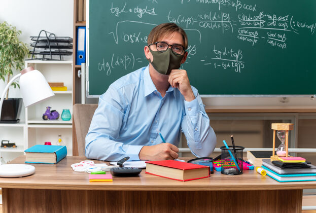 教室年轻的男老师戴着眼镜和面罩 严肃地看着摄像机 坐在课桌旁 教室的黑板前放着书和笔记桌子保护笔记