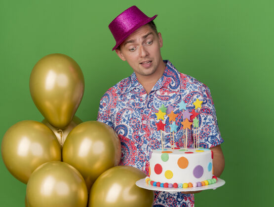 站着惊讶的年轻人戴着粉红色的帽子站在气球旁边 拿着蛋糕 看着绿色的隔离粉色穿上派对