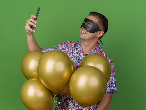 抱着高兴的年轻人戴着化装眼罩站在气球中间 拿着手机看着绿色的隔离小伙子眼睛化装舞会