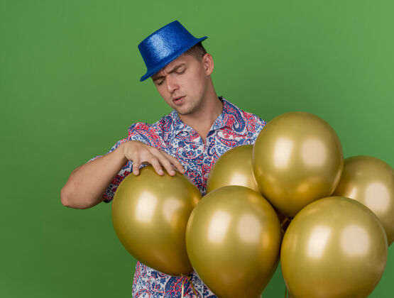 抓住体贴的年轻人戴着蓝色帽子站在气球后面 抓住了绿色的气球派对年轻小伙子