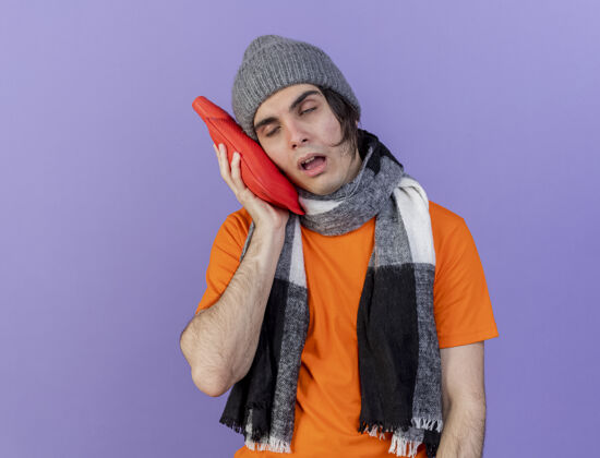 水体弱多病的年轻人戴着冬天的帽子 戴着围巾 把热水袋放在脸颊上 隔离在紫色上穿疾病紫色