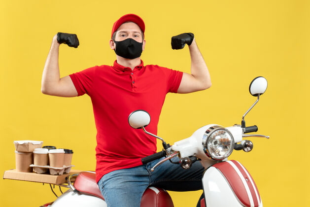 帽子俯视图自豪的年轻人穿着红色上衣 戴着帽子手套 戴着医用口罩 坐在滑板车上传递命令 展示他的肌肉肌肉男性摩托车
