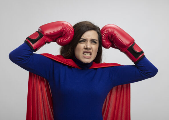 头恼怒的女超人穿着红色斗篷戴拳击手套把拳头放在头上隔离在白墙上姿势表情女孩