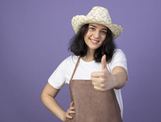 脸微笑着的年轻黑发女园丁穿着制服戴着园艺帽大拇指孤立地站在紫色的墙上拇指表情市民