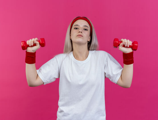 人自信的年轻运动女性戴着背带 戴着头带 手腕上拿着哑铃 隔离在粉红色的墙上哑铃戴年轻