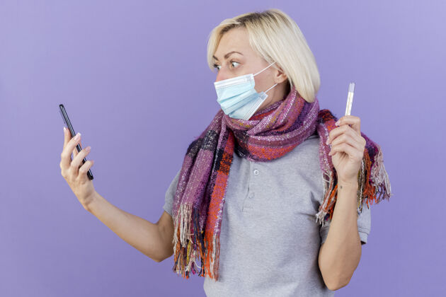 疾病一个戴着医用面罩和围巾的年轻金发病女人拿着温度计 看着隔离在紫色墙上的电话医疗金发印象