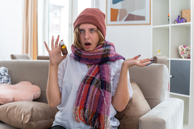 不适不健康的年轻女子戴着帽子 脖子上围着暖和的围巾 感觉不舒服 生病 患感冒和流感 手里拿着药瓶 坐在客厅里的沙发上 感到紧张和烦恼流感病药