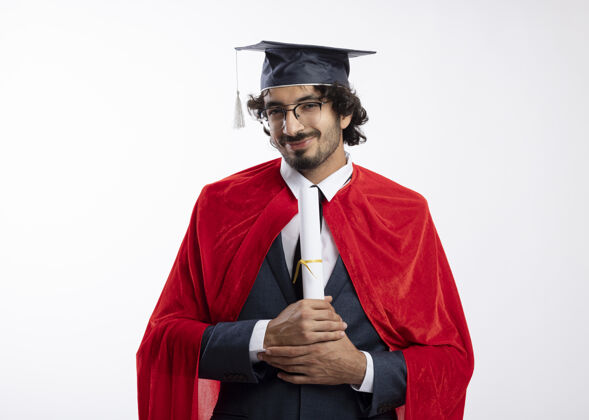 人戴着眼镜 身穿红色斗篷 头戴毕业帽的年轻超级英雄拿着毕业证书 望着隔离在白墙上的前方表情毕业人