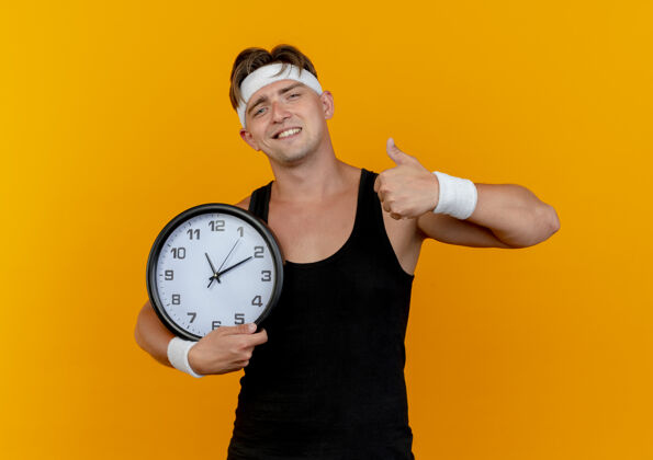 人高兴的年轻英俊的运动型男子戴着头带和腕带拿着时钟和显示拇指孤立在橙色的墙上人橙色请