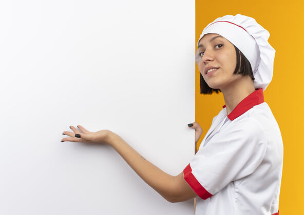 表情身着厨师制服 面带微笑的年轻女厨师站在白色墙壁旁 手拿着并指着隔离在橙色墙壁上的厨房女性脸手