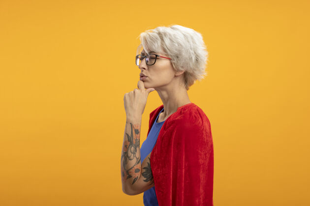 姿势戴着光学眼镜的戴着红色斗篷的困惑女超人站在一旁 手放在隔离在橙色墙上的下巴上表情斯拉夫人