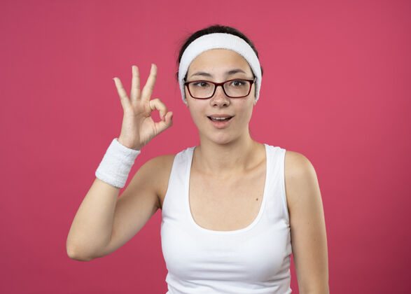人兴奋的年轻运动女性戴着眼镜戴着头带和腕带手势ok手势孤立在粉红色的墙上眼镜头带兴奋