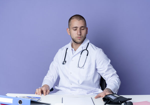 衣服神情严肃的年轻男医生穿着医用长袍 手持听诊器坐在办公桌旁 手放在办公桌上 低头看着隔离在紫色墙上的办公桌下市民年轻人