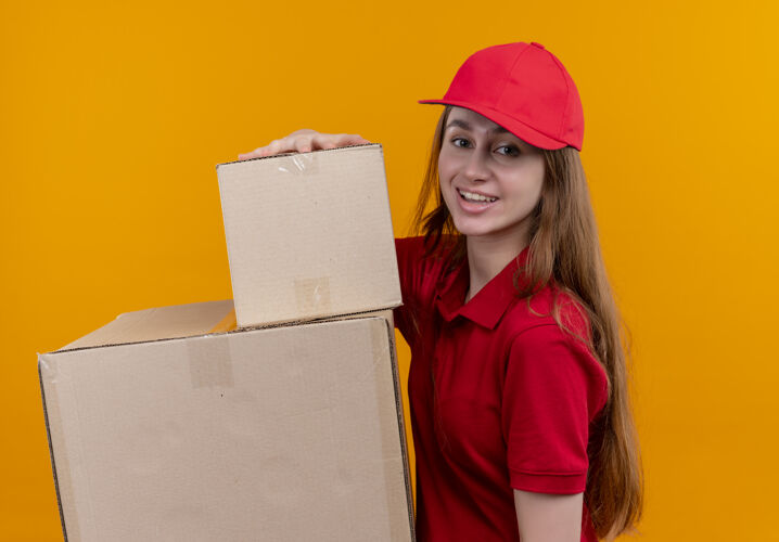 微笑微笑着的年轻送货女孩穿着红色制服拿着箱子站在孤立的橙色空间的侧视图递送年轻女孩