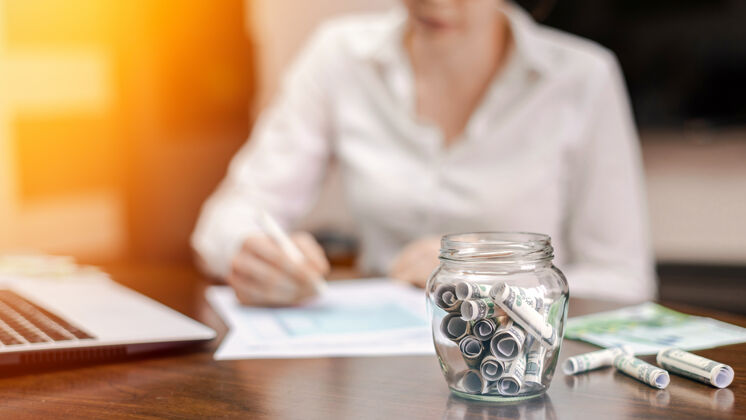 投资桌子上放着一个装满钞票的罐子笔记本电脑 纸张 背景是女人金融美元滚