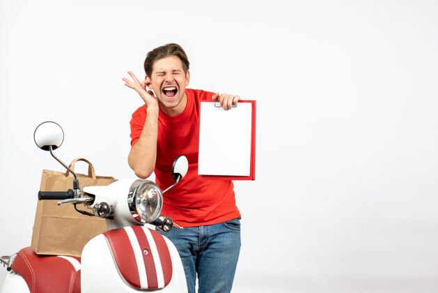 人年轻的情绪紧张的送货员穿着红色制服 站在滑板车旁 在白墙上展示文件合上了他的一只耳朵交付家伙快乐情绪