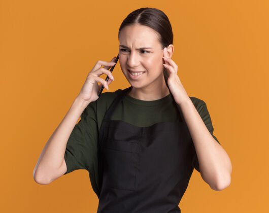 黑发恼怒的年轻黑发女理发师在制服闭上耳朵 在电话里看着橙色的一面制服关闭电话