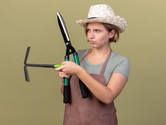 壁板自信的年轻斯拉夫女园丁戴着园艺帽手持园艺剪刀和锄头耙望着橄榄绿的一面剪刀橄榄帽子