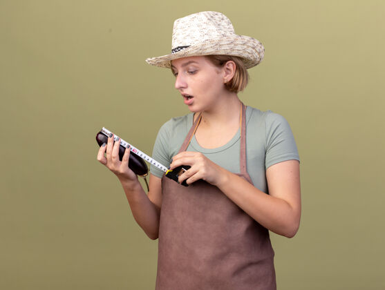 测量令人印象深刻的年轻斯拉夫女园丁戴着园艺帽测量茄子与卷尺在橄榄绿雌斯拉夫幼