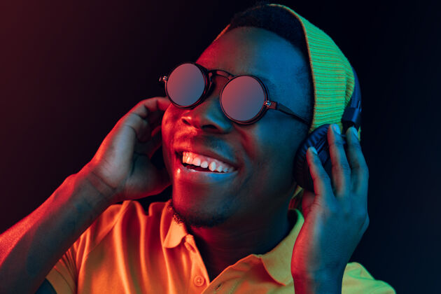 牙买加年轻帅气快乐的时髦男人在霓虹灯下用耳机在黑工作室听音乐迪斯科舞厅 夜总会 嘻哈风格 积极的情绪 面部表情 舞蹈概念音乐非洲耳机