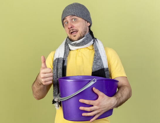 橄榄快乐的年轻金发病斯拉夫男子戴冬季帽子和围巾竖起大拇指 并持有橄榄绿墙上的复制空间隔离塑料桶喜悦举行疾病